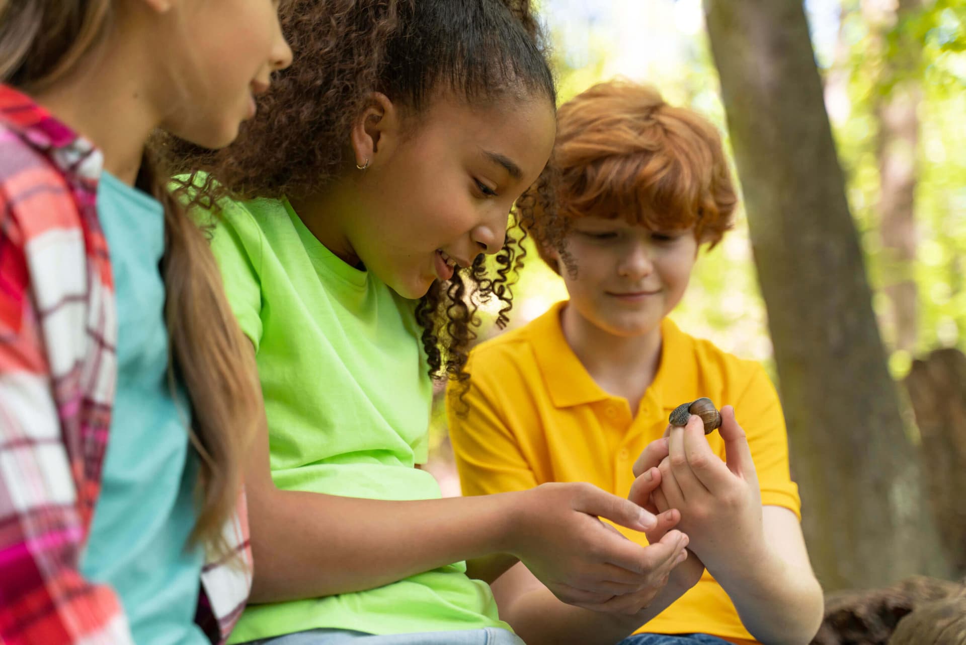 Three children outdoors in the woods looking closely at a snail resting on a boy’s hand