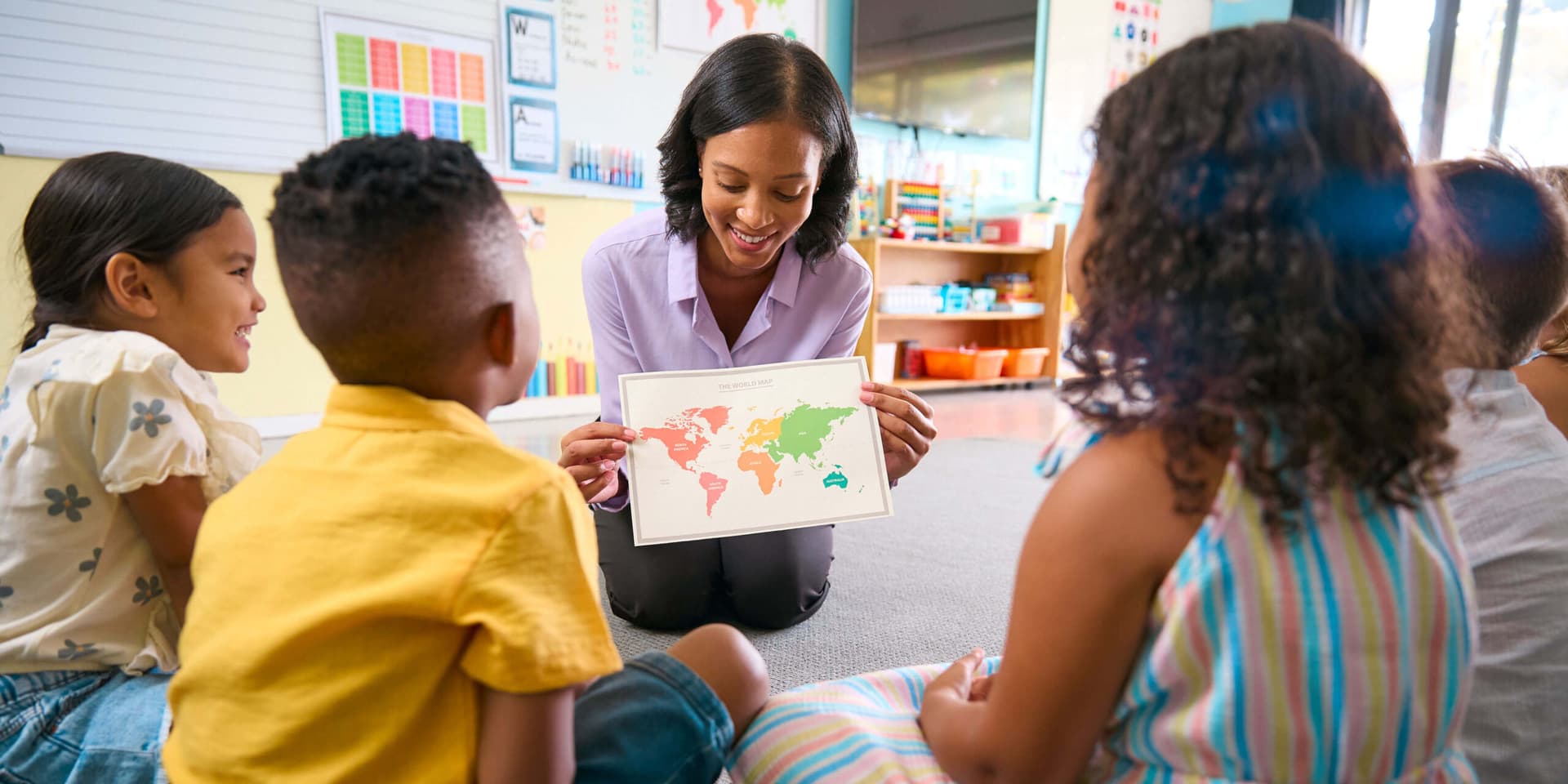 A teacher holding up a world map for a group of children seated on the classroom floor