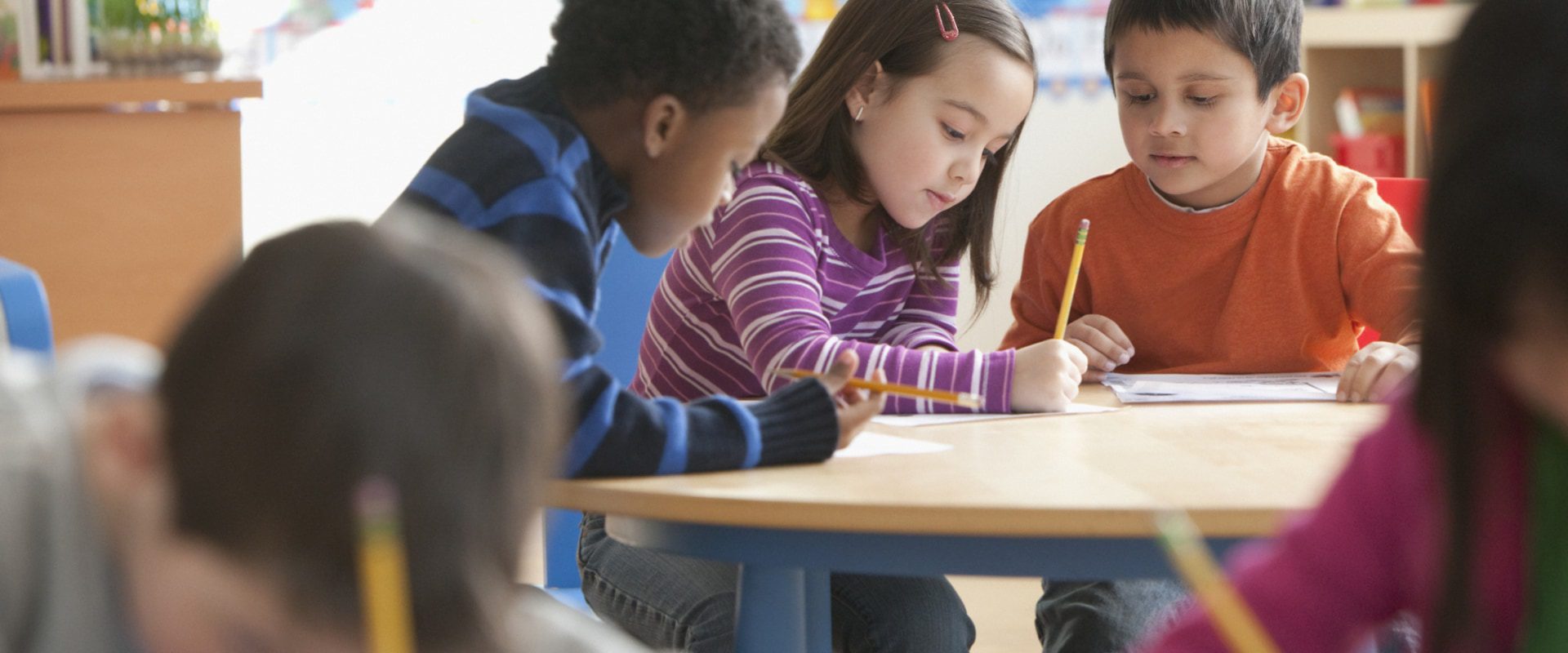Three children at a classroom table with paper and pencil.